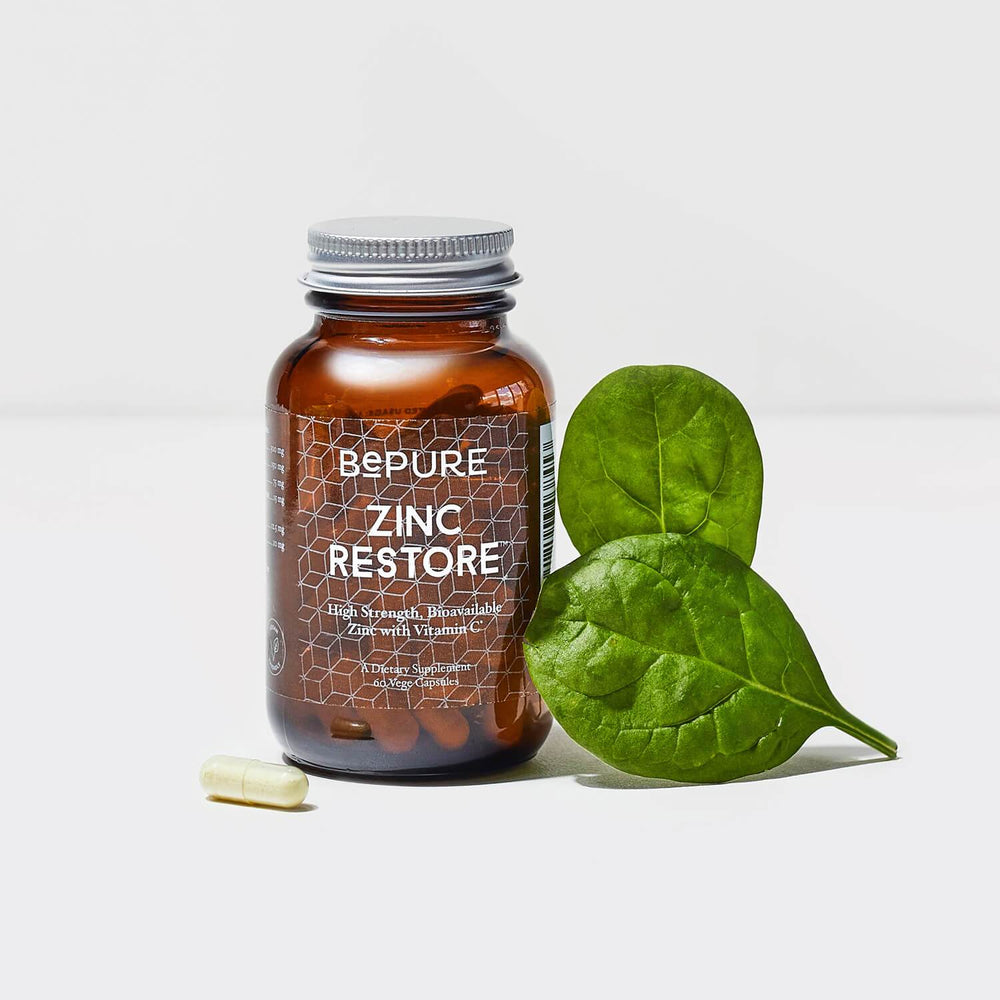 Photo of BePure Zinc Restore bottle (amber glass with clear label and white writing, silver looking screw cap lid on top), a capsule on the bottom left next to it and on the right 2 staged green leaves